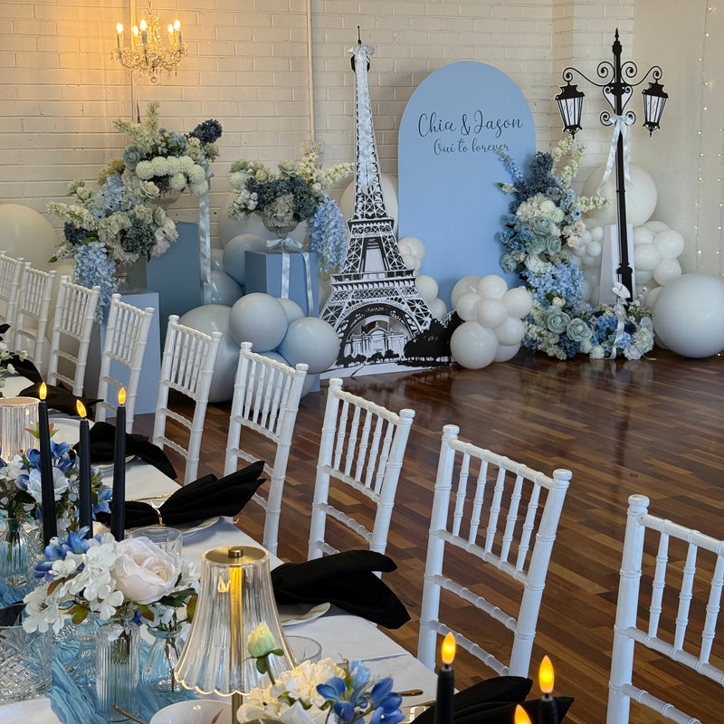 A decorated event space with a Parisian-themed setup, featuring a large light blue backdrop with a black Eiffel Tower silhouette, white and blue floral arrangements, white balloons, and rows of white chairs around tables with black napkins and lit candles.