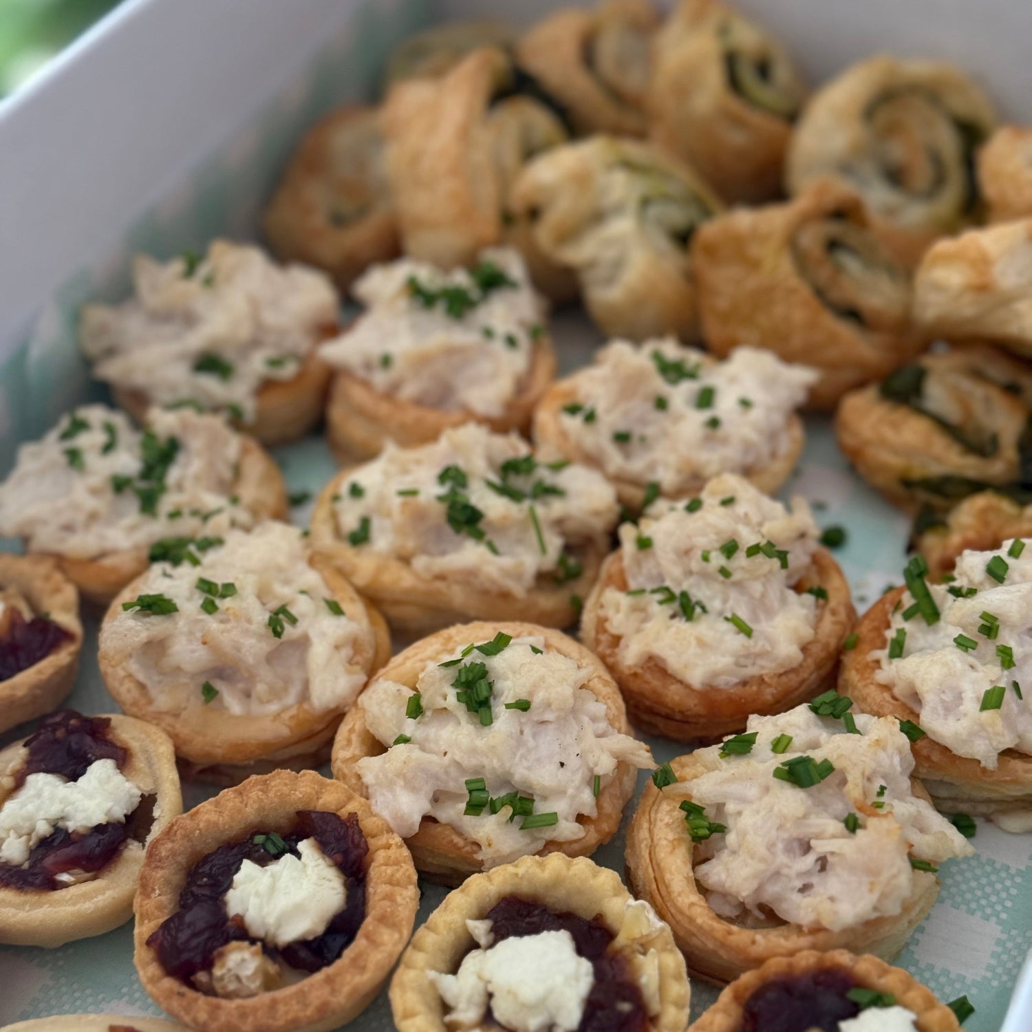 A tray of assorted appetizers including stuffed pastry cups, small tartlets with cream and red jam, and rolled pinwheel appetizers, arranged in a white serving box.