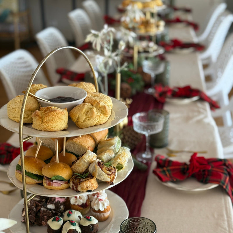 A three-tiered white serving stand filled with assorted pastries, scones, sandwiches, and cupcakes, set on a decorated table with red napkins, glassware, and a long table in the background.