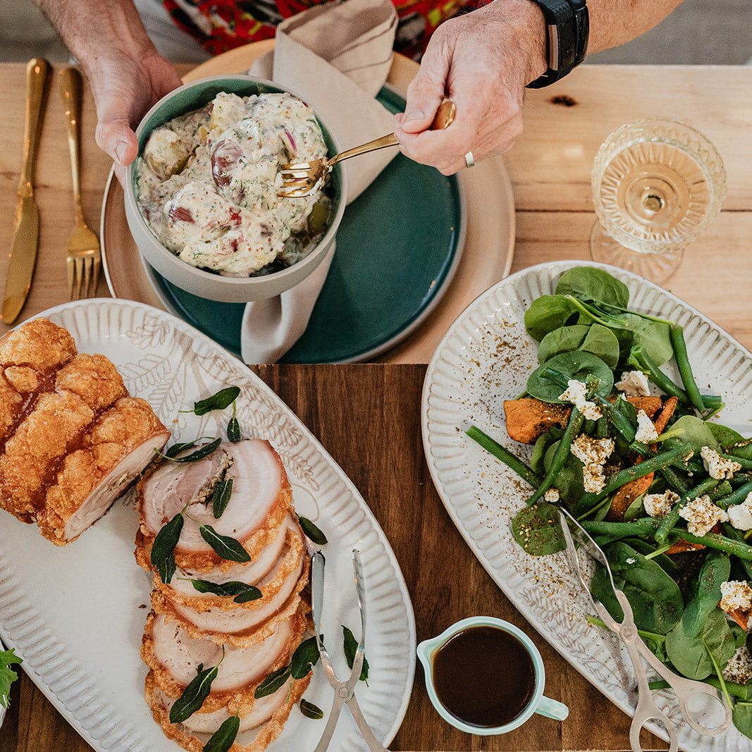 A styled holiday dining table featuring a large platter of sliced turkey with sage, a green salad with spinach, feta, and carrots, a bowl of creamy stuffing, and bread on white oval platters, with hands serving food in the background.