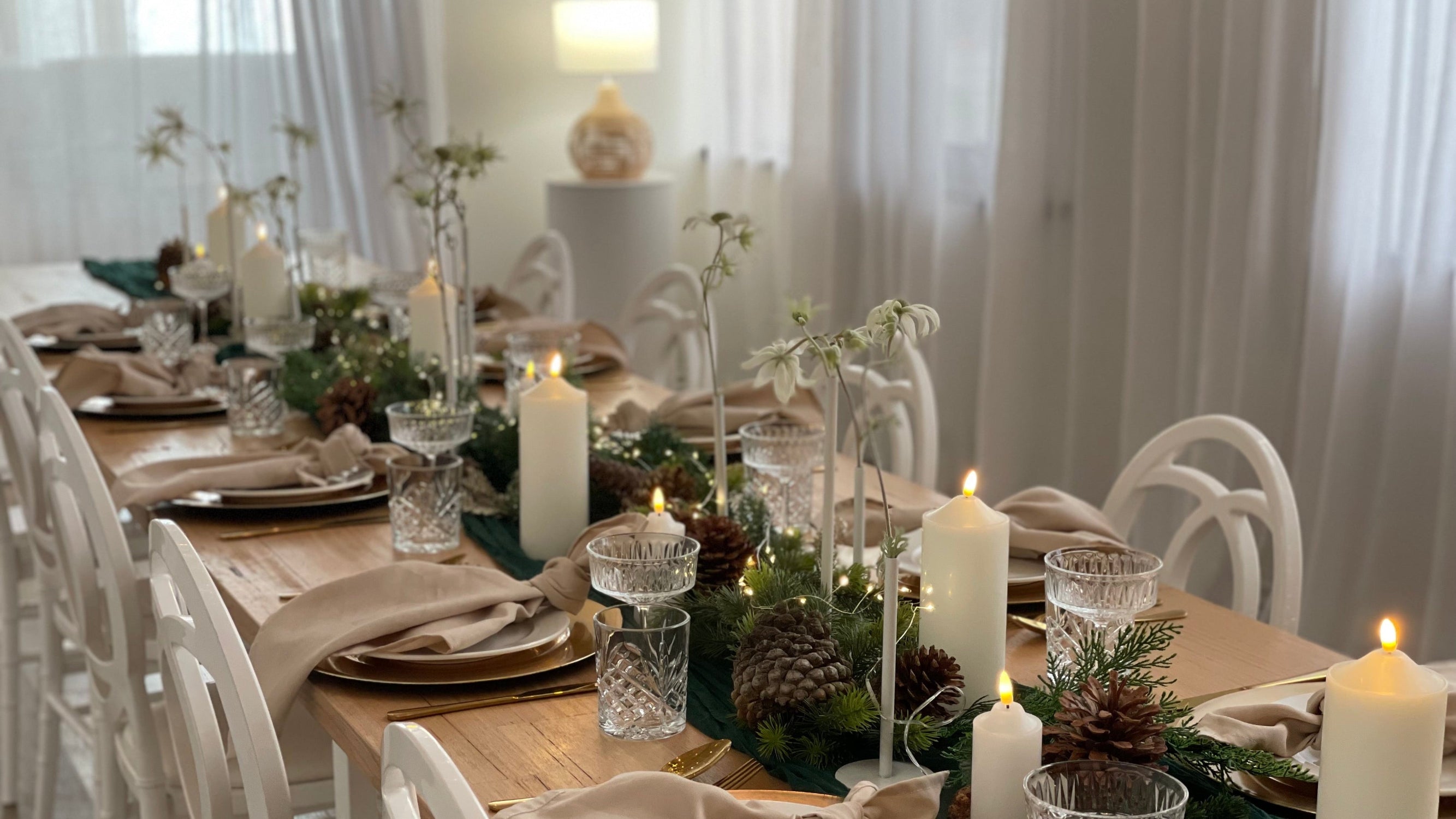 A beautifully set dining table for a holiday gathering, featuring white chairs, gold-rimmed plates, beige napkins, clear glass candleholders, and green garlands with pinecones and lit white pillar candles in a bright, softly lit dining room.