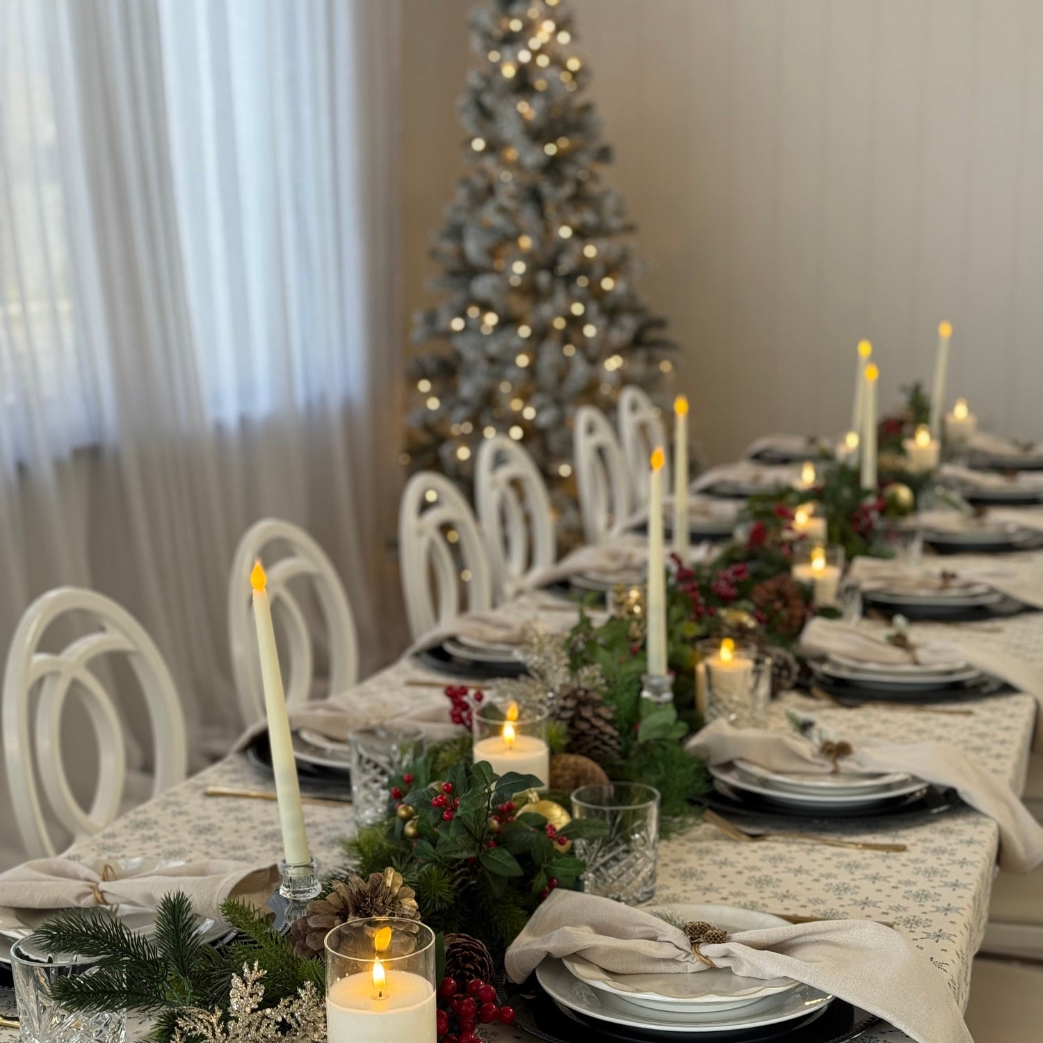 A long dining table set for a holiday feast with a snowflake-patterned tablecloth, white plates, folded cream napkins, lit taper candles, and festive green garlands with red berries. White chairs surround the table, and a softly lit Christmas tree stands in the background.