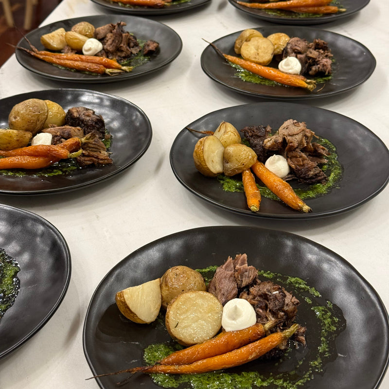 Multiple black oval plates arranged on a white table, each plated with cooked meat, whole potatoes, carrots, and green sauce.