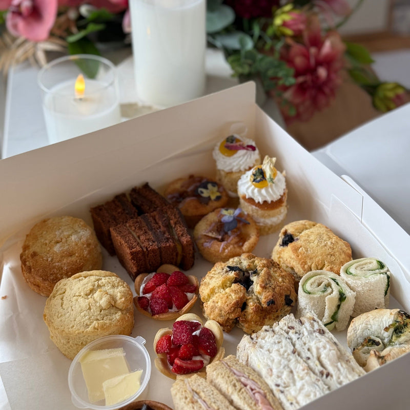 A white cardboard box filled with assorted finger foods and sandwiches, photographed on a table with lit candles and flowers in the background.