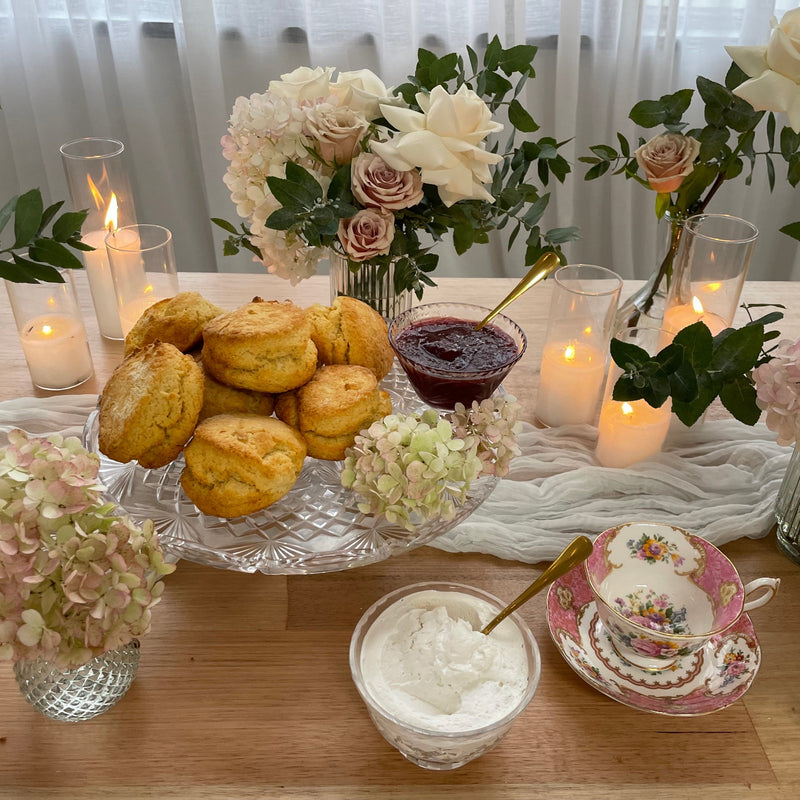 A styled breakfast or tea spread on a wooden table with golden scones on a glass platter, a bowl of jam, a bowl of whipped cream, a floral teacup and saucer, and multiple flower arrangements with lit candles in the background.