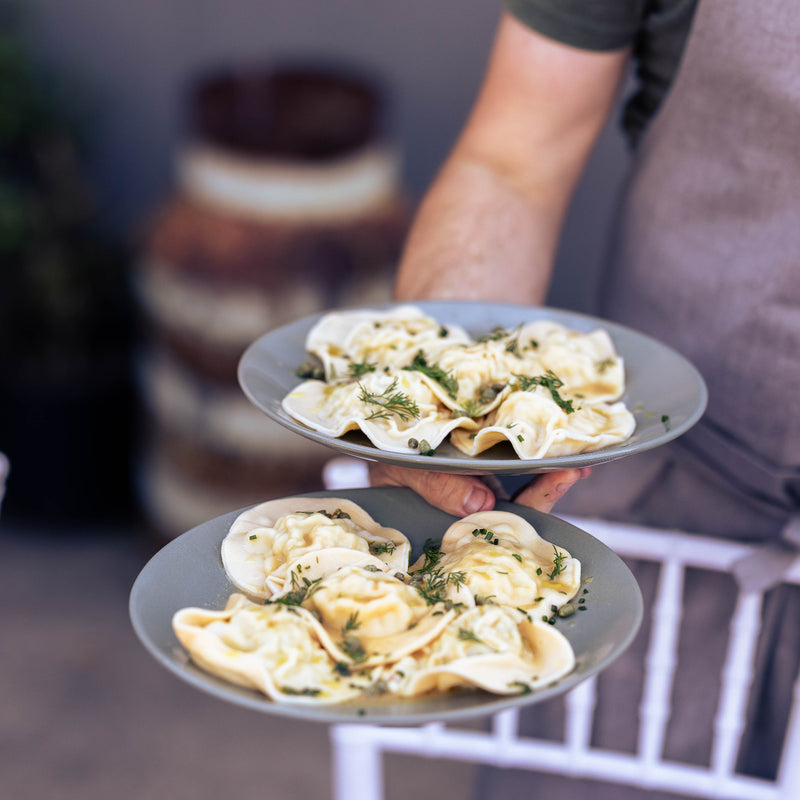 A person wearing a gray apron holds two gray plates of stuffed pasta shells topped with green herbs in a softly blurred kitchen or dining setting.