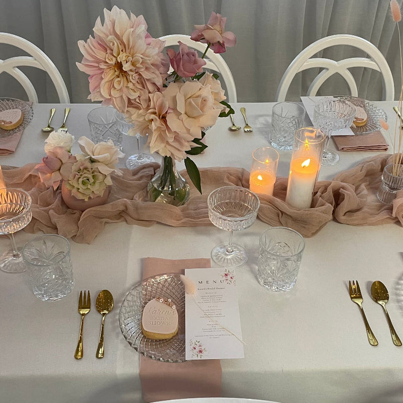 A styled wedding reception table setting with a blush pink and beige drape runner, white tablecloth, clear glass plates and goblets, gold cutlery, and a central floral centerpiece with lit candles.