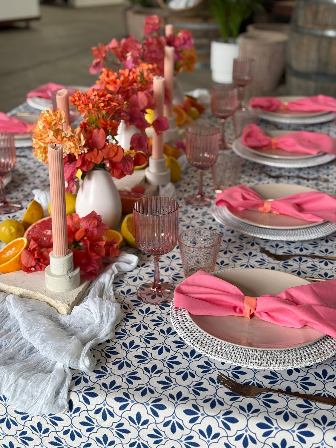 A styled dining table setting with a blue and white patterned tablecloth, pink glassware, white plates, woven placemats, and bright floral centerpieces with pink napkins. European summer