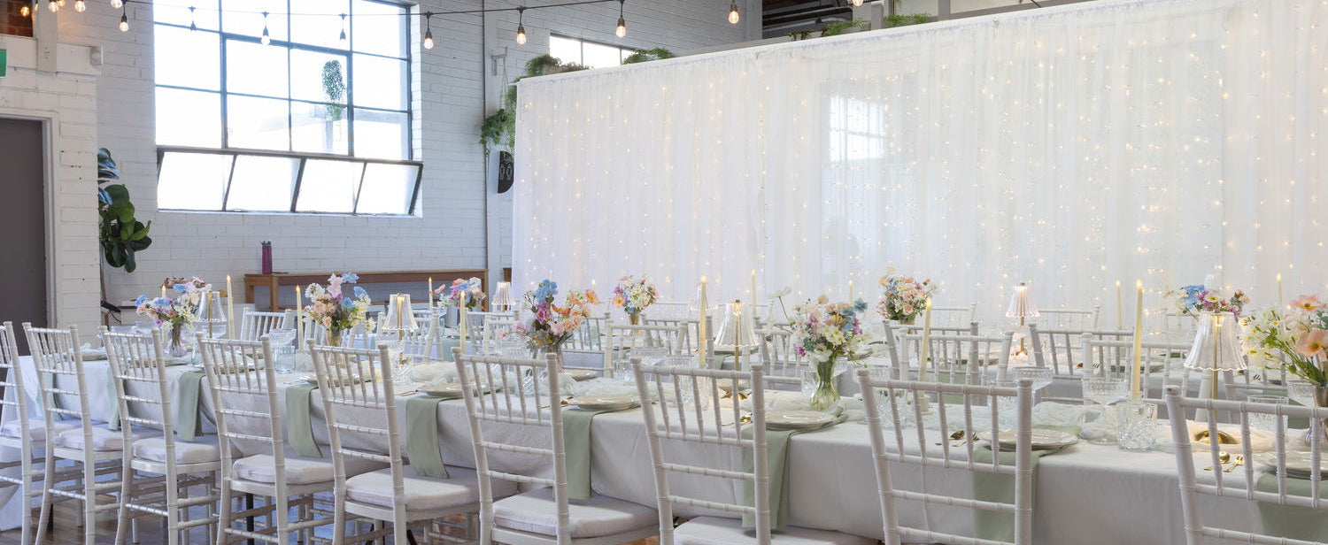 A long dining table set in a bright event hall with white chairs, white tablecloths, floral center pieces, and a large white draped backdrop covered in string lights.