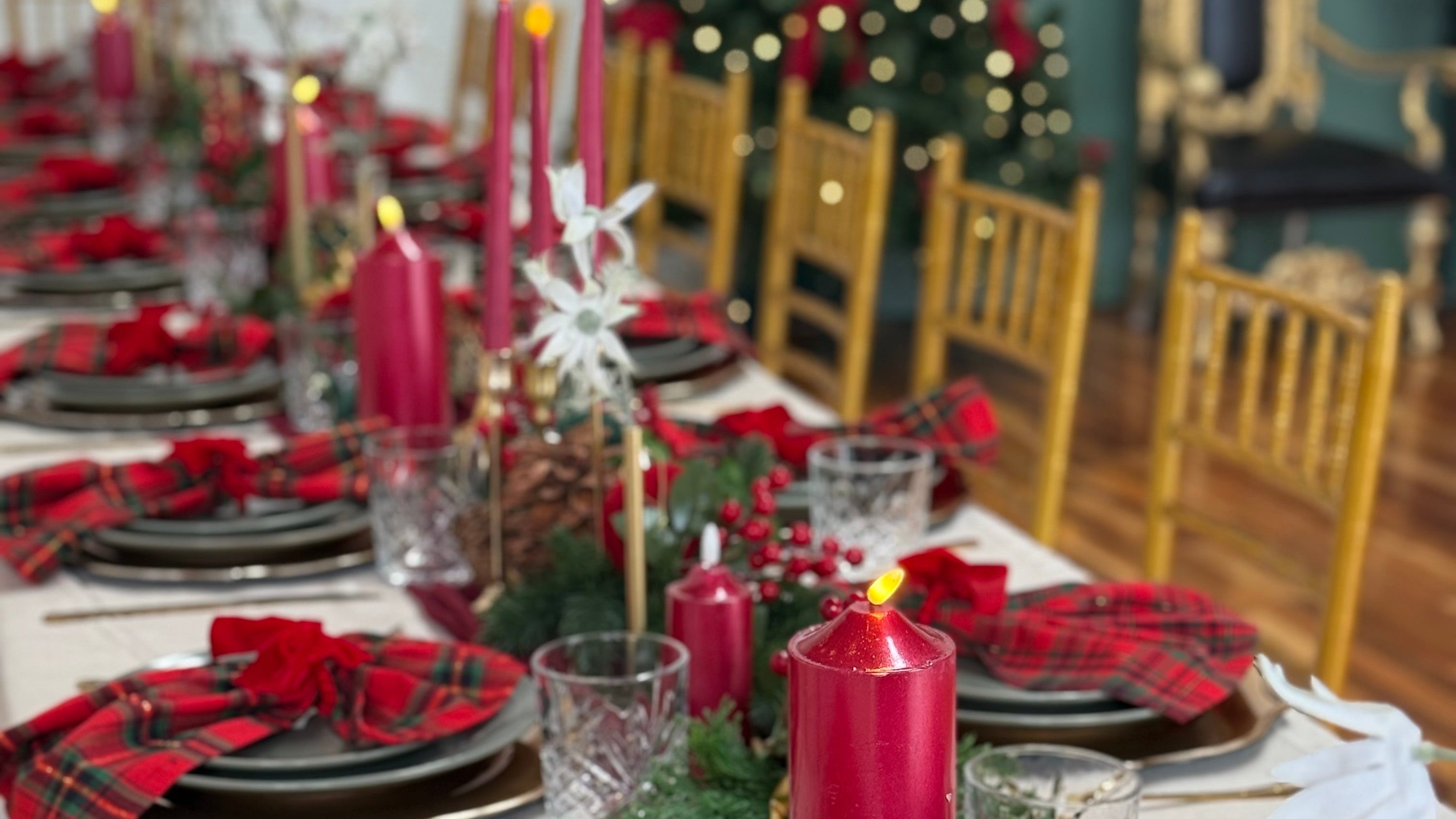A festive holiday table setting with red plaid napkins, red taper candles, clear glassware, and pinecones on a white tablecloth, with a decorated Christmas tree in the background.