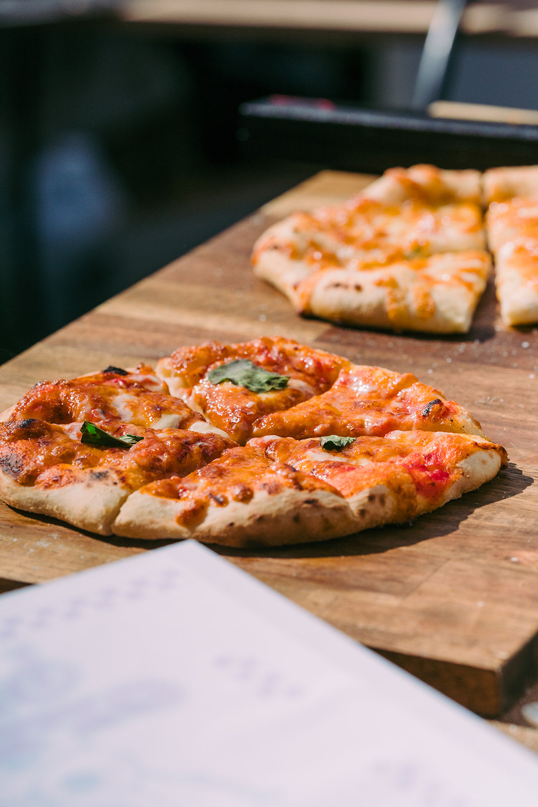 Two pizzas on a wooden cutting board with a blurred background