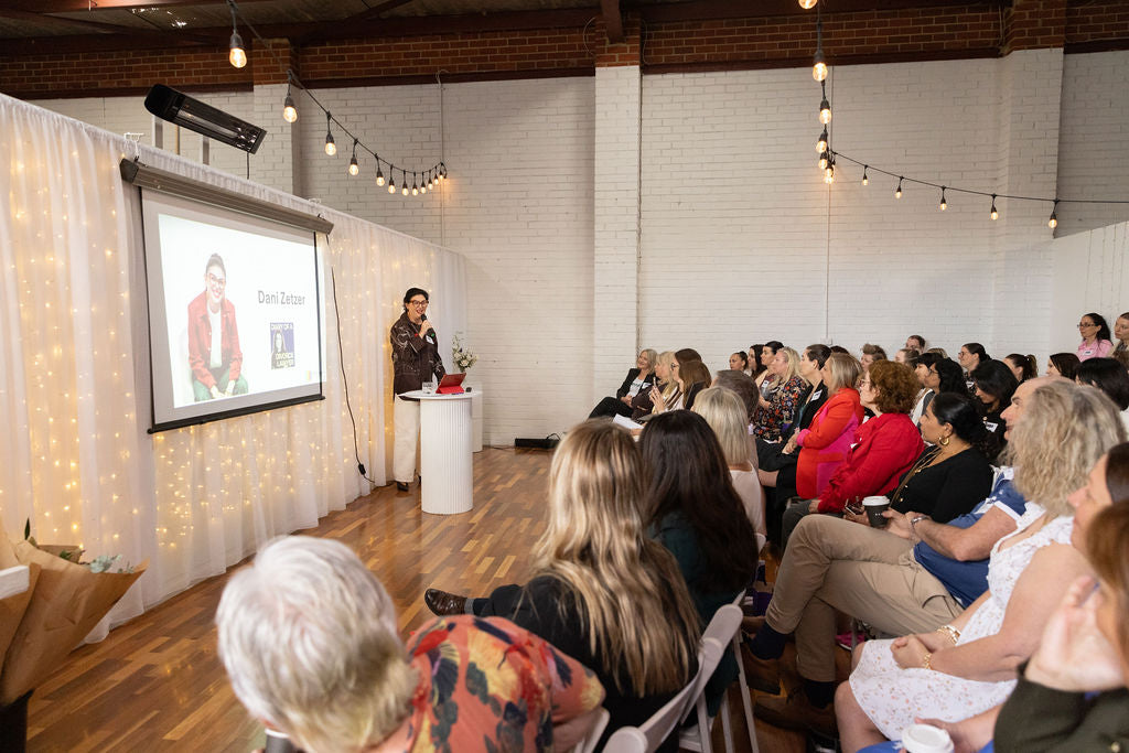 Person giving a presentation to an audience in a room with a projector screen and string lights.