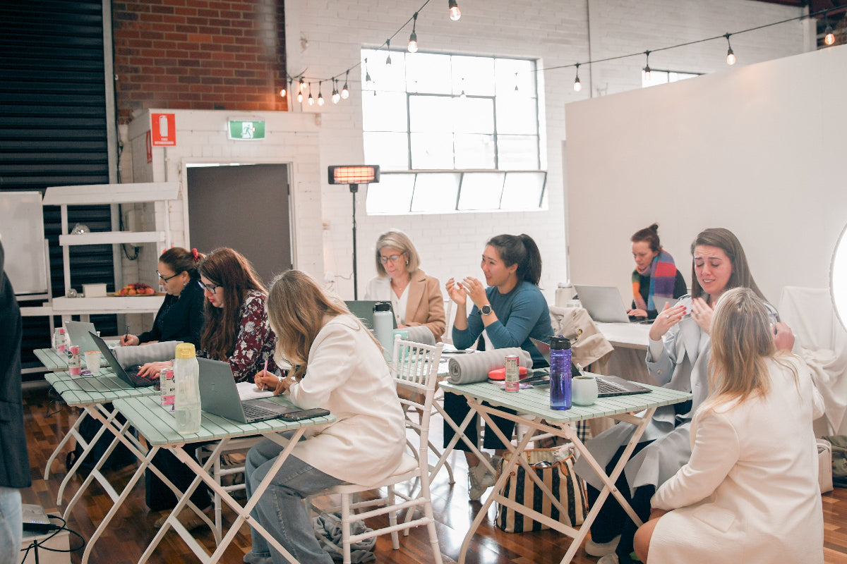 A group of women collaborating in a bright, modern office space with white folding tables, laptops, and large windows.