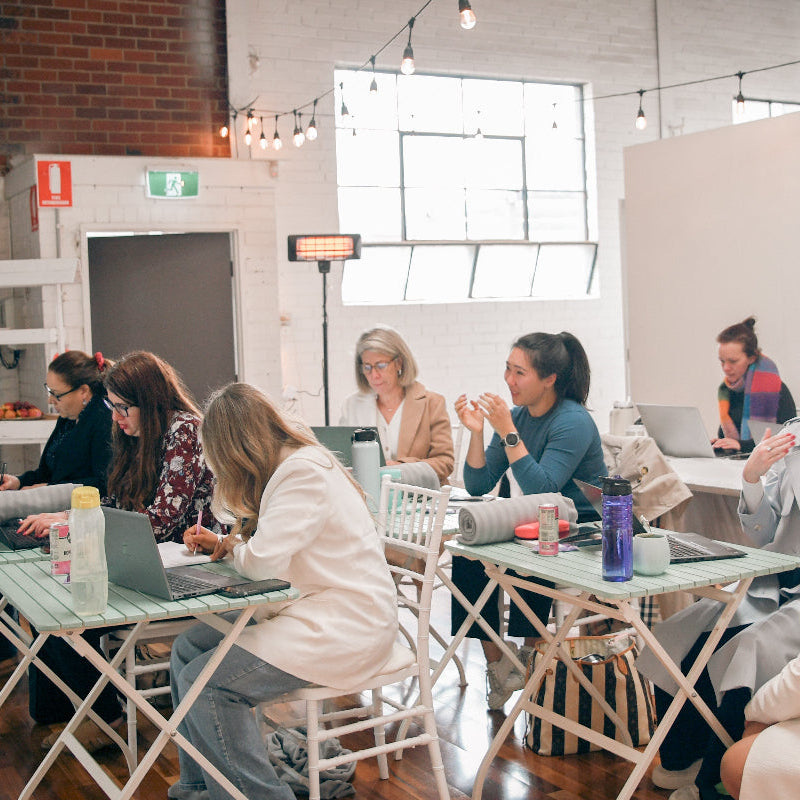 A group of women collaborating in a bright, modern office space with white folding tables, laptops, and large windows.