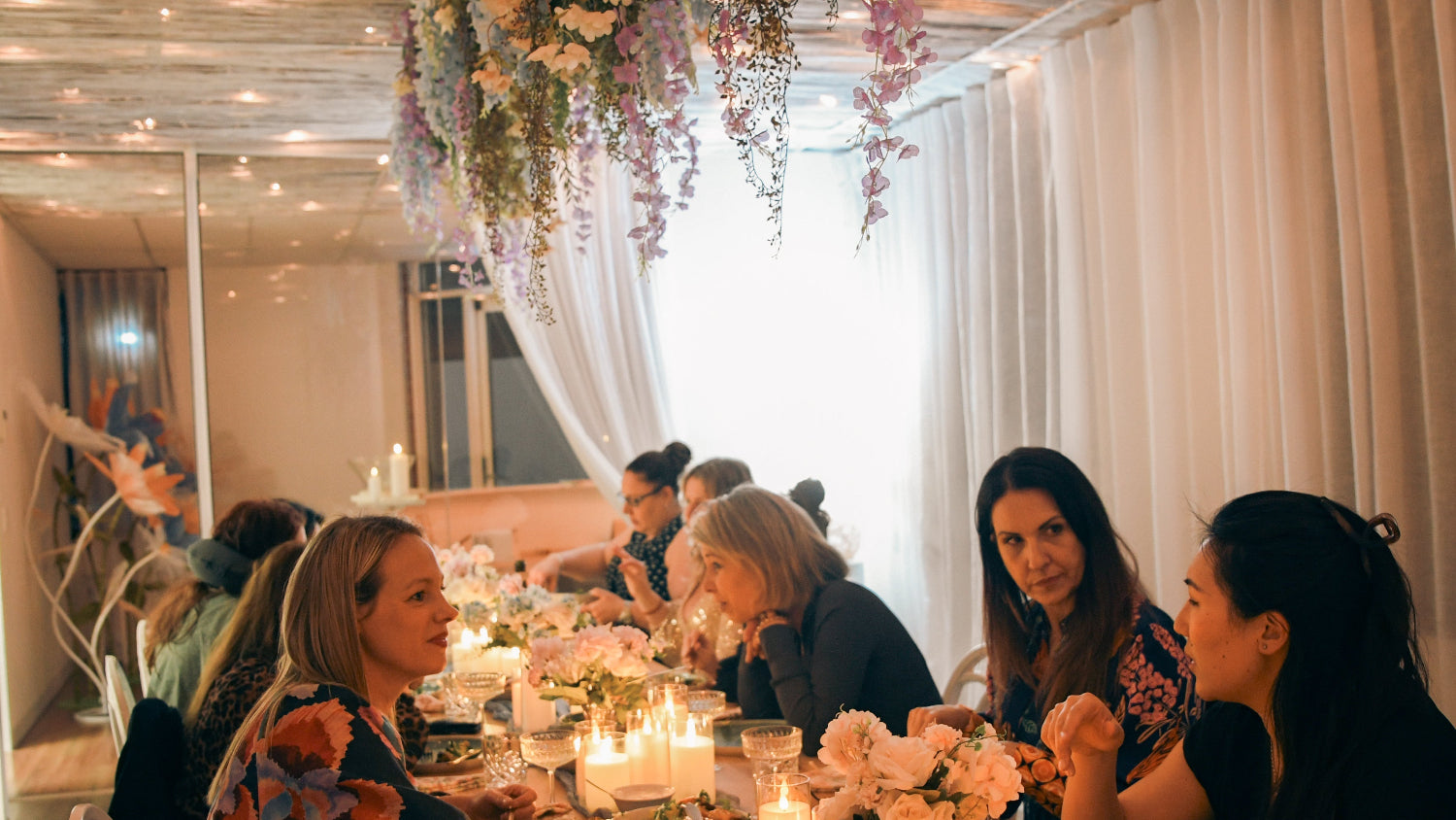 A warm, elegant indoor gathering scene with several women seated around a long table decorated with candles, flowers, and string lights. White drapes hang in the background, and floral arrangements are suspended from the ceiling.