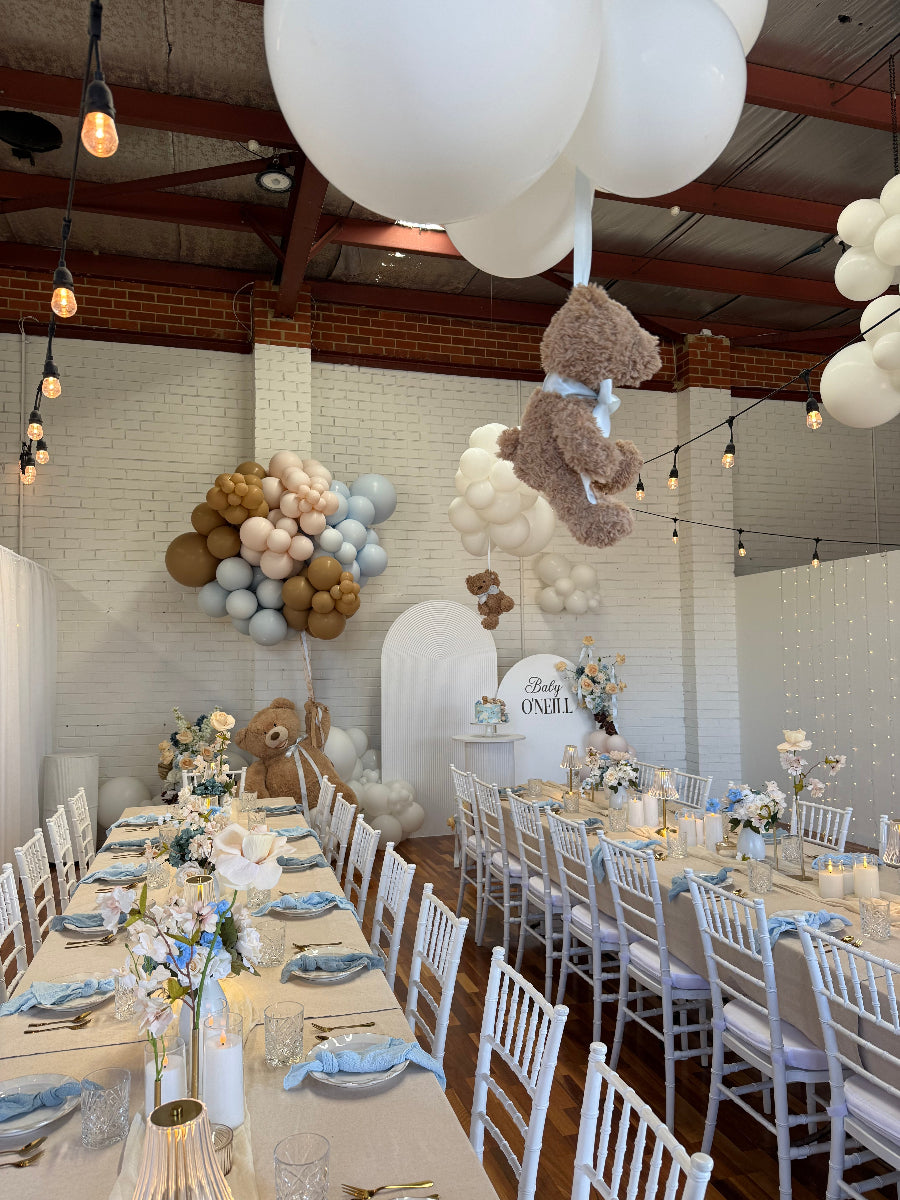 A decorated event space with long tables set for guests, white chairs, balloon arrangements in white, beige, and light blue, and several plush teddy bears hanging and sitting on the floor. 